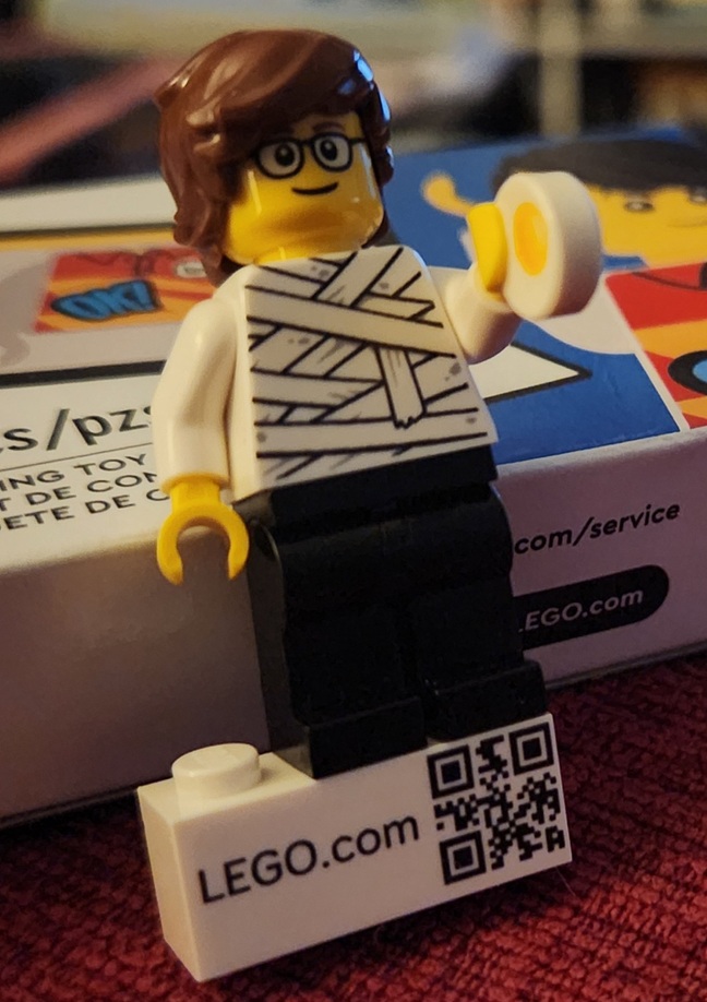 Lego minifigure: messy brown hair, glasses, mummy top, black pants, holding 1x1 flat egg and standing on a LEGO.com brick